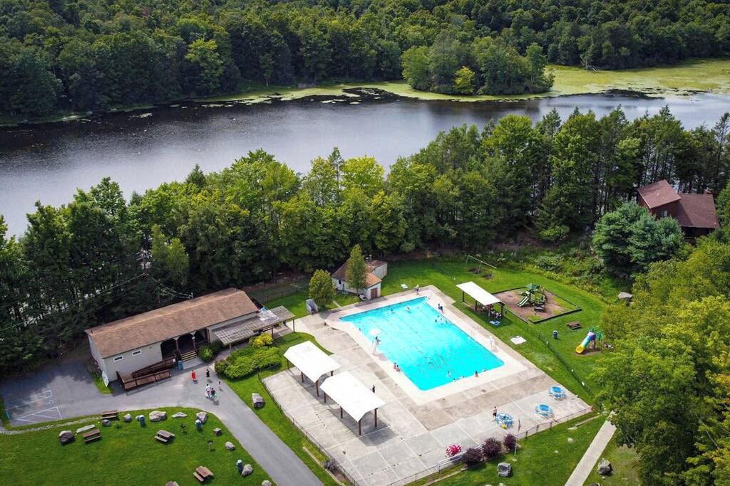 A swimming pool surrounded by a covered deck area, a small building, and a parking lot.