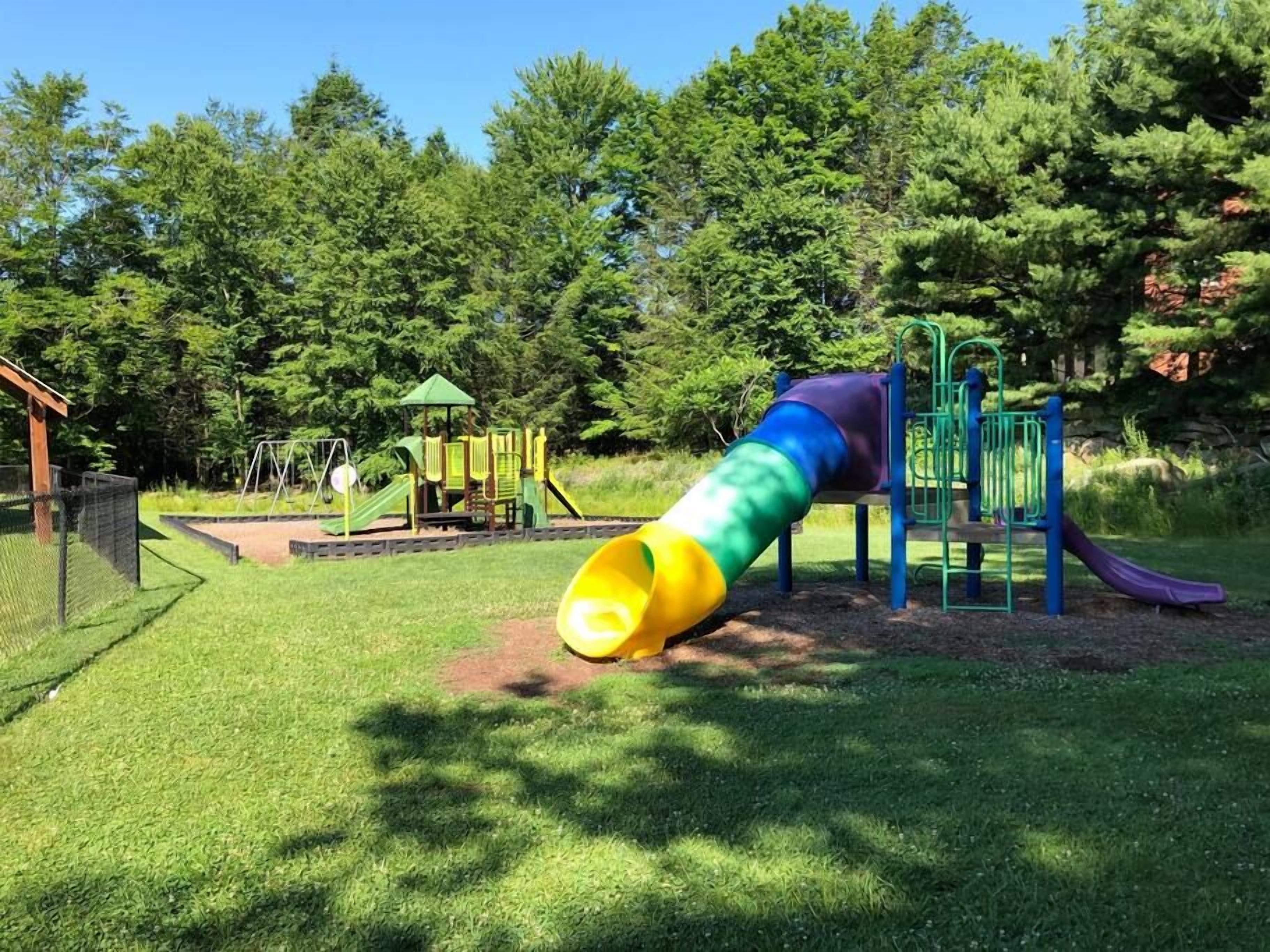 A playground with a slide and climbing structures, surrounded by grass and trees.