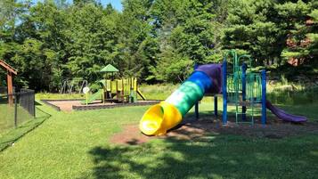 A playground with a slide and climbing structures, surrounded by grass and trees.