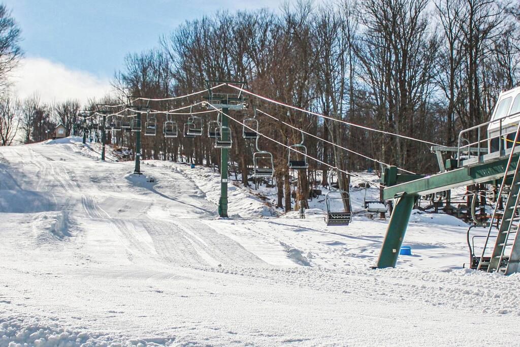 Een besneeuwd gebied met een skilift, stoelen en kabels, omgeven door bomen en een heldere lucht.
