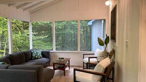 Una sala de estar acogedora con un sofá gris, una mesa de centro de madera y una silla de madera. Amplias ventanas ofrecen vistas al bosque.