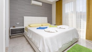 Ein modernes Schlafzimmer mit einem weißen Bett, einem Nachttisch, einer Wand-Klimaanlage und einem großen Fenster mit durchsichtigen Vorhängen.