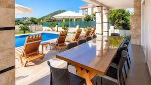 Un espace aménagé au bord de la piscine, avec une table et des chaises en bois, une vue dégagée sur la piscine et un bâtiment en arrière-plan.