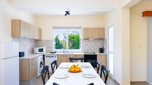 A modern kitchen with a dining table set for four, a washing machine, and a window with a view of greenery. - Sun Beach Villa Thio (Poli Crysochous)