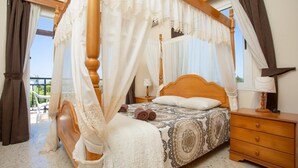 A four-poster bed with a canopy, a bedside table, a lamp, and a view of the outdoors. - Sun Beach Villa Thio (Poli Crysochous)