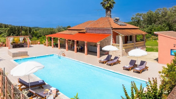 A pool area with lounge chairs and umbrellas, a covered seating area, and a stone building with a tiled roof.