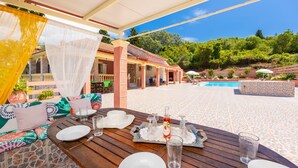 An outdoor dining area with a pool, covered seating, and a view of the pool and greenery.