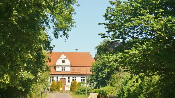 A white building with a red roof and a central entrance, surrounded by lush greenery. - Einfach Urlaub auf Rügen (Groß Schoritz)