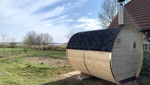 A wooden sauna with a sloped roof and a chimney, situated in a grassy area with a view of a lake and trees in the background. - Einfach Urlaub auf Rügen (Groß Schoritz)