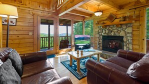 A cozy cabin interior with a stone fireplace, leather sofas, and a large window overlooking a scenic landscape. - Free Attractions | Secluded, New Hot Tub w/ Views, Fireplace, Pet-Friendly (Gatlinburg)