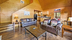A spacious log cabin interior with a living area featuring two leather sofas, a coffee table, and a kitchen area with wooden cabinets and a refrigerator. - Free Attraction Passes | Firepit, Theater, Pool Table, Hot Tub, Private (Sevierville)