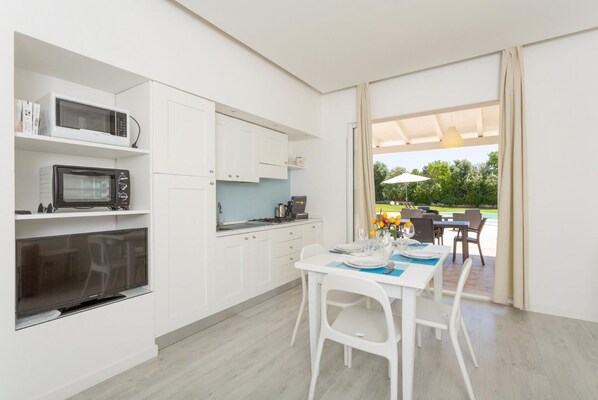 A modern kitchen with white cabinets, a microwave, and a built-in oven. There is a dining area with a table set for two, and a view of an outdoor patio with a pool and umbrellas. - Villino Malva Large Private Pool, A/C, WiFi (Scicli)