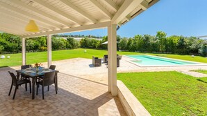 Eine überdachte Terrasse mit Tisch und Stühlen, ein Schwimmbad und eine gepflegte Rasenfläche.