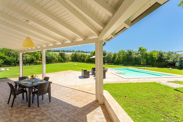 Eine überdachte Terrasse mit Tisch und Stühlen, ein Schwimmbad und eine gepflegte Rasenfläche.