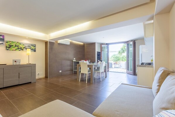 Una sala de estar moderna con un sofá, un comedor con mesa y sillas, y una cocina visible a través de una puerta abierta.