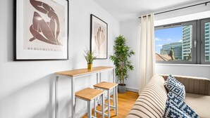 Una sala de estar moderna con un sofá, una pequeña mesa de comedor con taburetes, dos obras de arte enmarcadas y un ventanal con vistas a la ciudad.