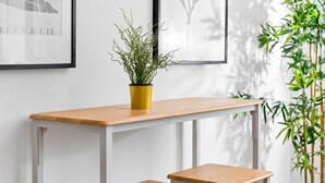 Habitación minimalista con pared blanca, una mesa de madera con dos bancos, una maceta amarilla con plantas verdes y obras de arte enmarcadas.