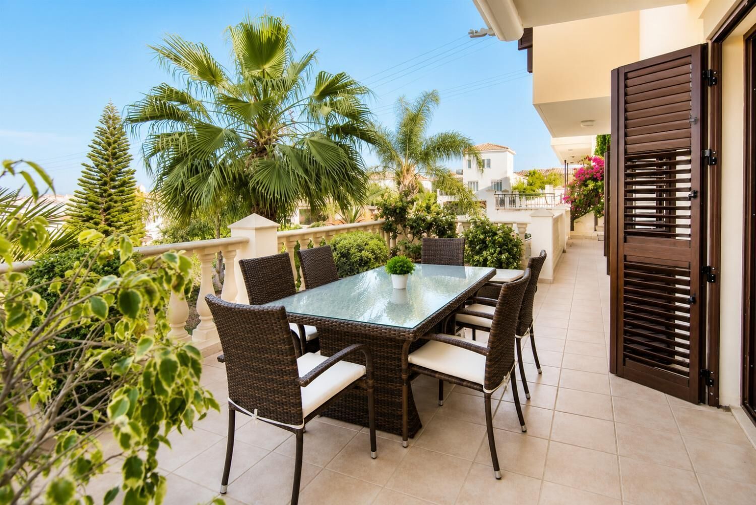 Eine Außenpatio mit einem Tisch aus Glas und Korbstühlen, umgeben von üppiger Vegetation und einer Palme.