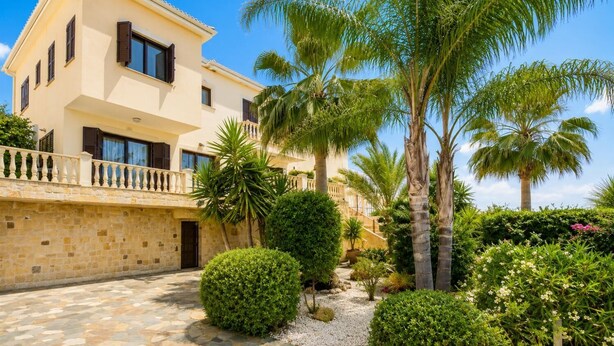 A two-story building with a stone facade, a balcony with a balustrade, and a garden with palm trees and shrubs.