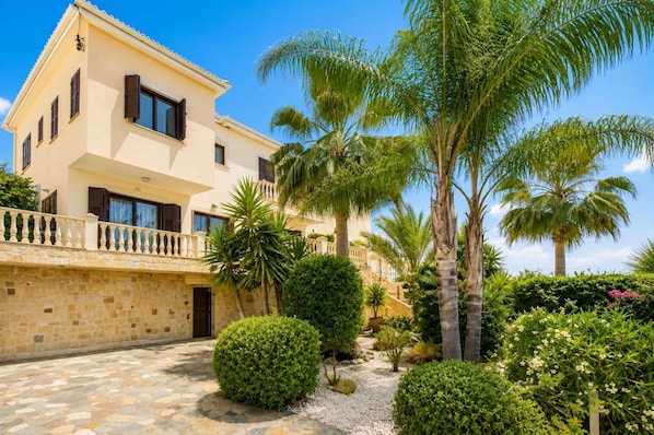 A two-story building with a stone facade, a balcony with a balustrade, and a garden with palm trees and shrubs.
