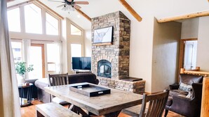 Un comedor con una mesa de madera, bancos y sillas. Hay una chimenea de piedra con un televisor montado encima.