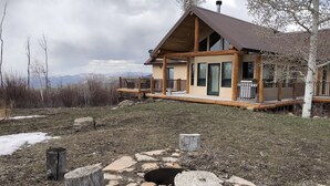 Una cabaña con una terraza de madera, un hogar de piedra y vistas a montañas al fondo.