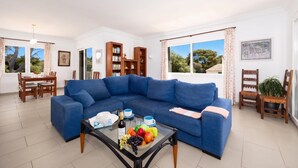 Un salon avec un canapé bleu, une table basse en verre sur laquelle se trouvent des fruits et une bouteille, un coin repas avec une table et des chaises, et une bibliothèque.
