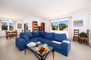 Een woonkamer met een blauwe bank, een glazen salontafel met fruit en een fles, een eethoek met een tafel en stoelen, en een boekenkast.