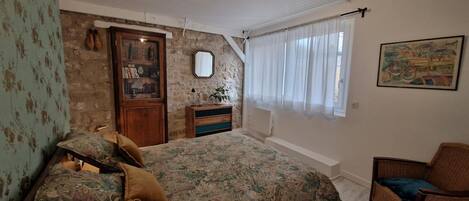 Une chambre à coucher avec un lit, une commode, une chaise, un miroir et une fenêtre avec des rideaux.