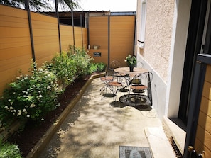 Um pequeno alpendre ao ar livre com uma mesa e cadeiras, cercado por um muro de tijolos baixo e diversas plantas.