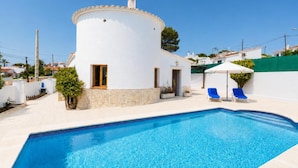 A white villa with a swimming pool, two blue lounge chairs, and a stone wall.