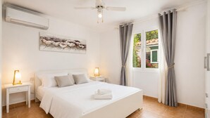 A bedroom with a bed, two bedside tables with lamps, a ceiling fan, a window with curtains, and an air conditioning unit.