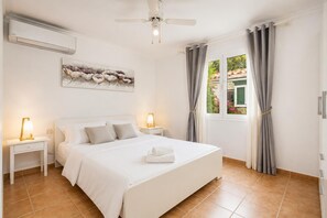 A bedroom with a bed, two bedside tables with lamps, a ceiling fan, a window with curtains, and an air conditioning unit.