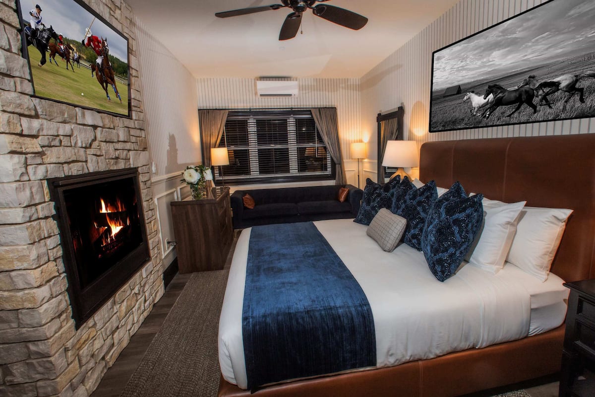 A bedroom with a stone fireplace, a large bed with blue bedding, a headboard, and a painting of horses on the wall.