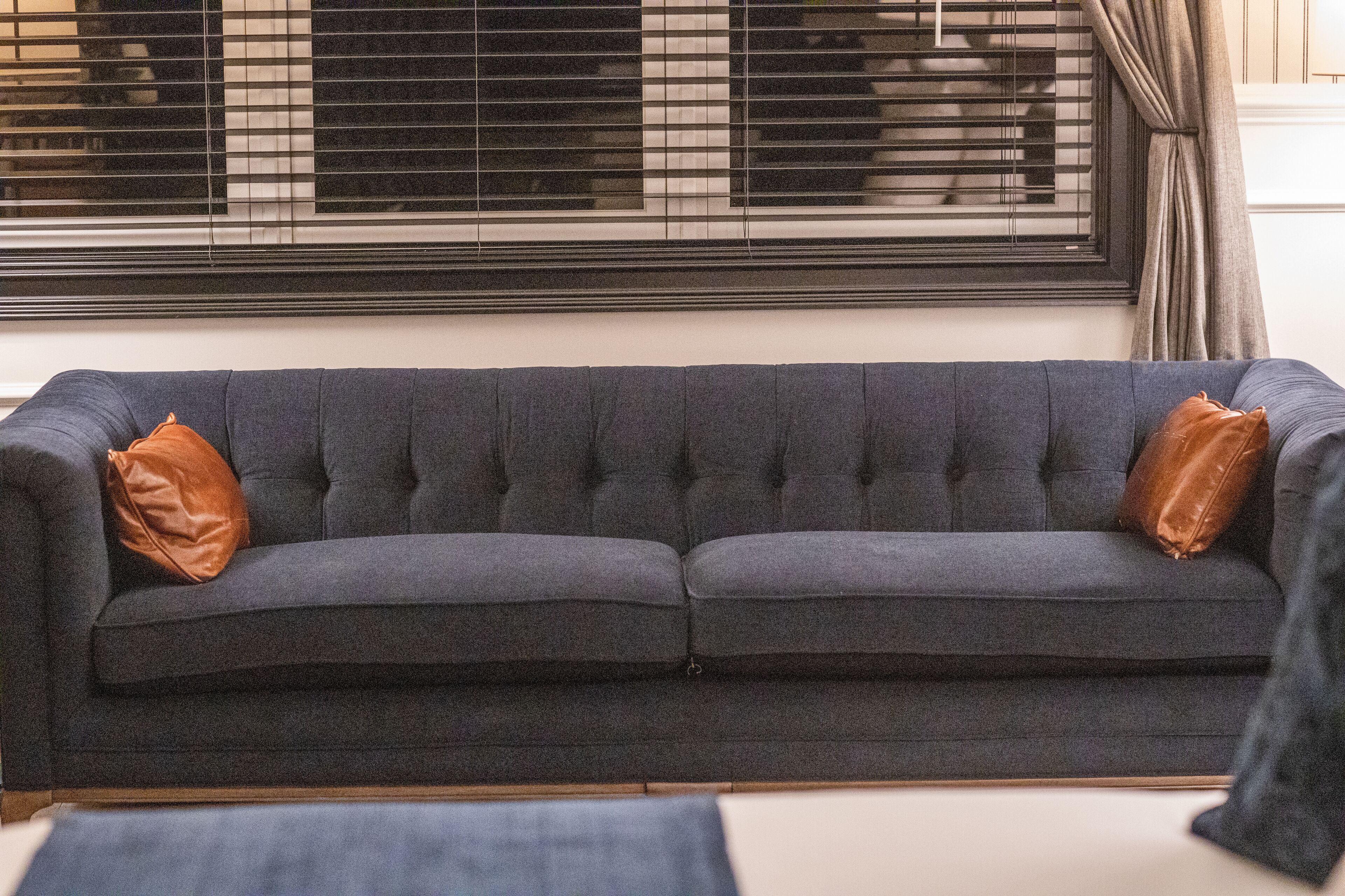 A dark grey sofa with two brown cushions, positioned in front of a window with blinds.