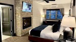 A hotel room with a stone fireplace, a bed with a blue and white duvet, a TV mounted on the wall, a ceiling fan, and a window with a view of trees. - The Belfry House (Williams Bay)