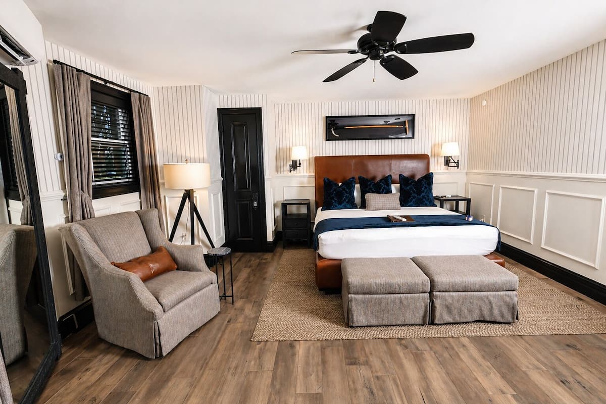 A hotel room with a bed, a chair, a desk, a standing lamp, a ceiling fan, and a window with blinds.