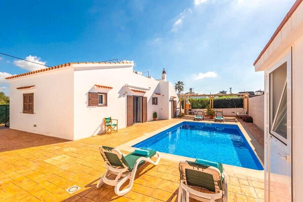 A white villa with a swimming pool, two lounge chairs, and a patio area.