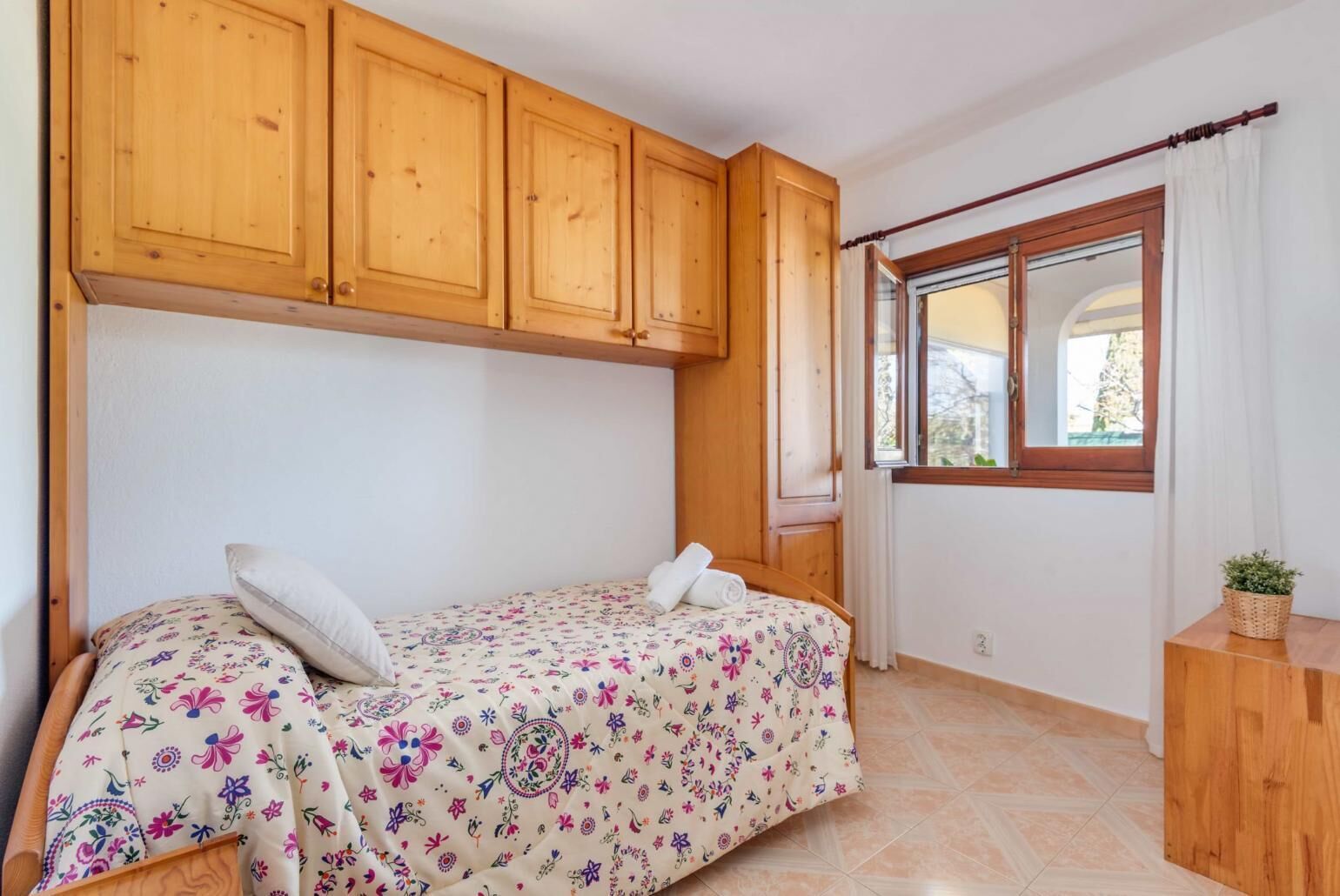 Ein Schlafzimmer mit einem Bett, Holzschränken, einem Fenster mit Vorhängen und einem kleinen Holztisch mit einer Pflanze.