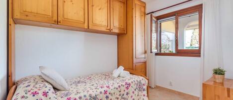 Ein Schlafzimmer mit einem Bett, Holzschränken, einem Fenster mit Vorhängen und einem kleinen Holztisch mit einer Pflanze.