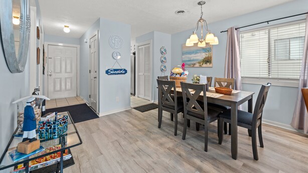 A dining area with a wooden table and chairs, a chandelier, and a view of the outside through a window with blinds. - Shipwatch Pointe I F117 Two Bedroom Condo First Floor Condo (Myrtle Beach)