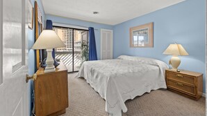 A bedroom with a bed, two bedside tables, a window with curtains, and a view of buildings outside. - Shipwatch Pointe I F117 Two Bedroom Condo First Floor Condo (Myrtle Beach)