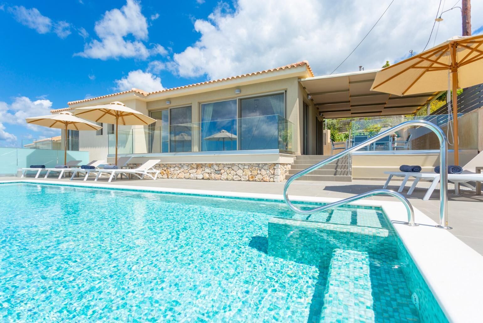 A swimming pool with clear blue water, surrounded by lounge chairs and umbrellas, and a building with large windows and a stone wall.