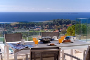 Ein Tisch ist reichhaltig mit Frühstück zubereitet und mit Orangensaftgläsern bestückt, mit Blick auf eine Küstenlandschaft mit Häusern und Bäumen.