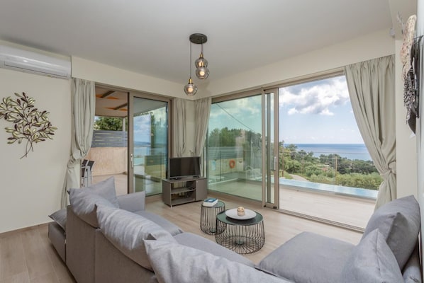 Un moderno salón con un sofá gris, un televisor y una mesita. Hay un balcón con vistas al mar.