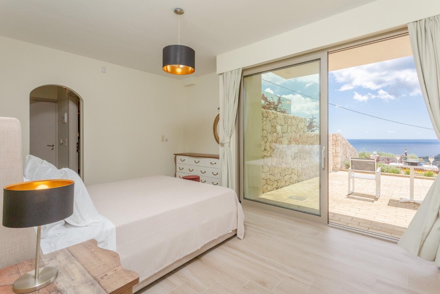 Een slaapkamer met een bed, een dressoir, een lamp en uitzicht op zee door een schuifdeur.