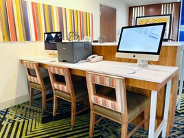 Un mostrador de recepción de hotel con dos computadoras Apple iMac, un teléfono de escritorio y un quiosco de registro. Hay tres sillas a rayas con asientos acolchados. El piso tiene una alfombra estampada.