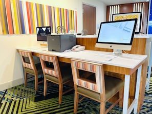 A hotel reception desk with two Apple iMac computers, a desk phone, and a check-in kiosk. There are three striped chairs with cushioned seats. The floor has a patterned carpet.