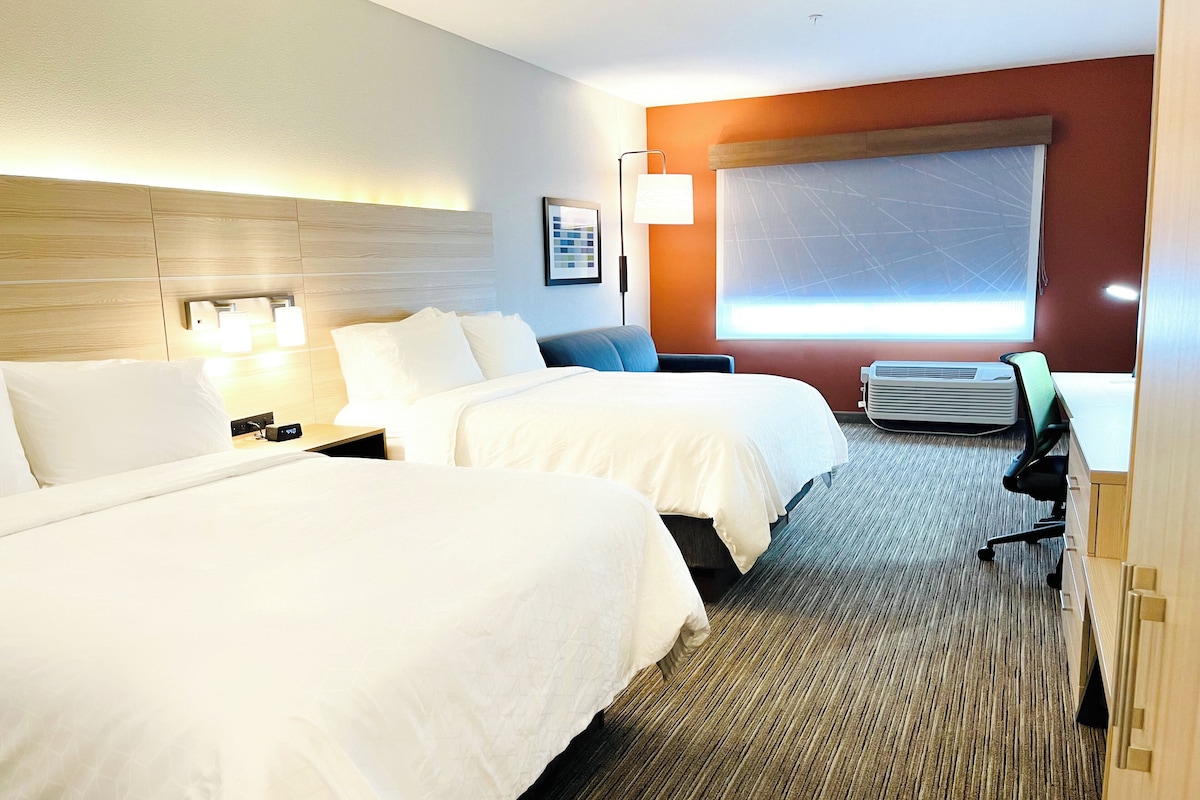 A hotel room with two beds, a desk, a chair, a lamp, and a large window with blinds.