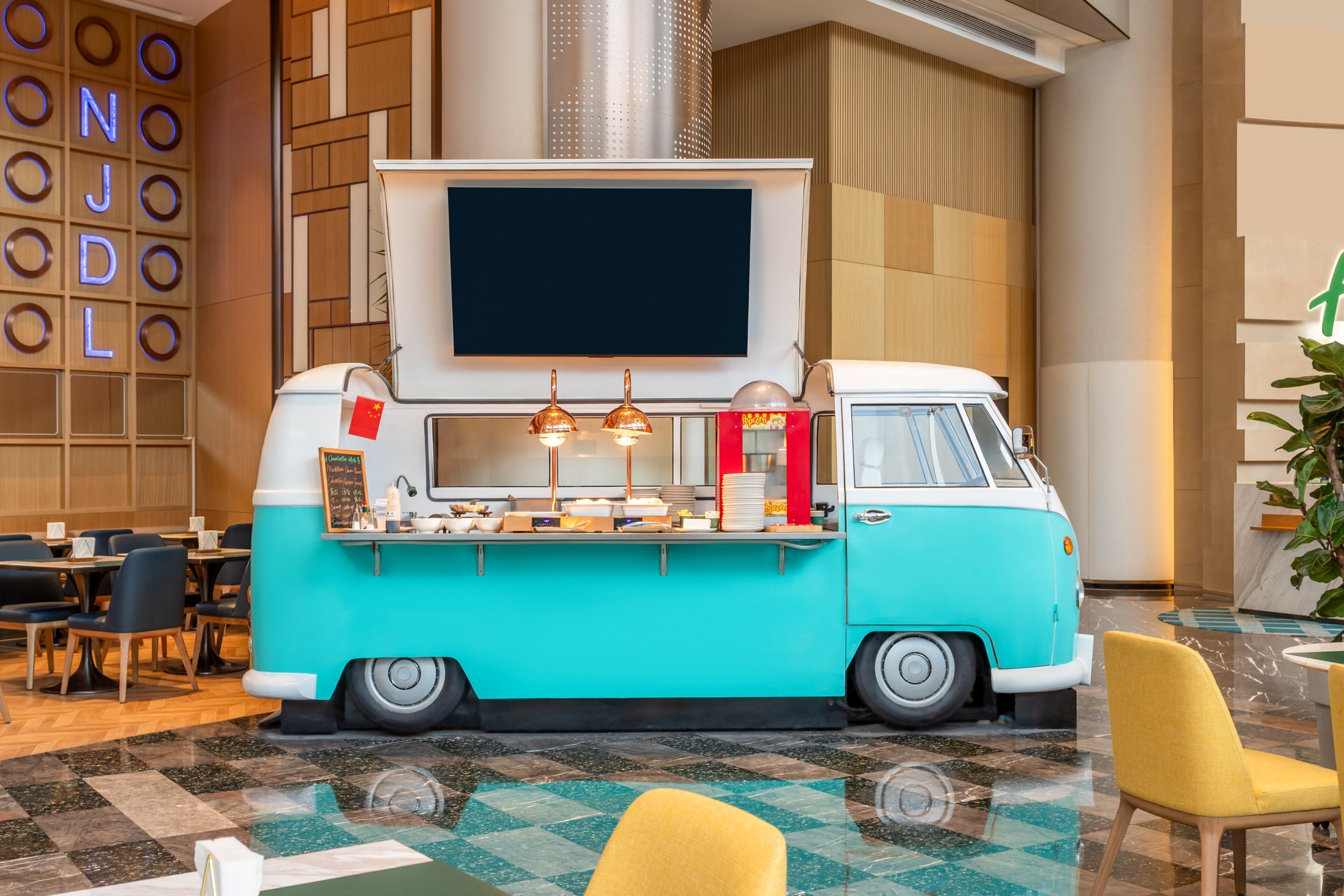 A retro-themed food truck in a modern hotel lobby with seating and a large screen.